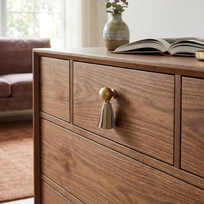 A close-up of a wooden dresser with a warm medium-brown finish, featuring a gold-toned tassel pull on the front drawer. 