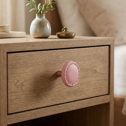 A close-up of a light oak nightstand featuring a round, pink faux-leather drawer knob with visible stitching. 