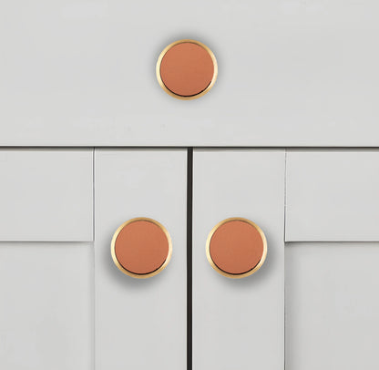 Leather knobs with gold trim mounted on a light gray cabinet, adding a warm, modern accent to the design by Mantara.