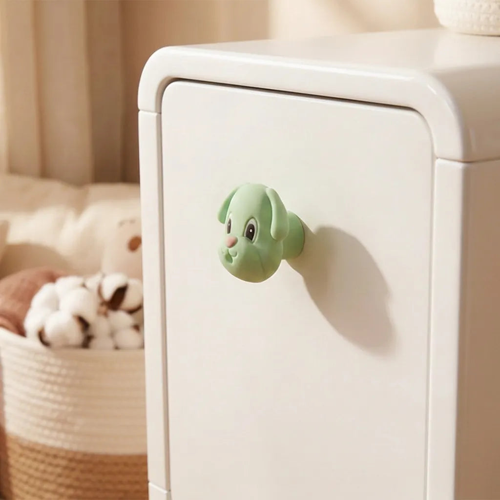 A small mint green dog-shaped drawer knob attached to a white cabinet door in a softly lit room, with a woven basket and plush toy blurred in the background.