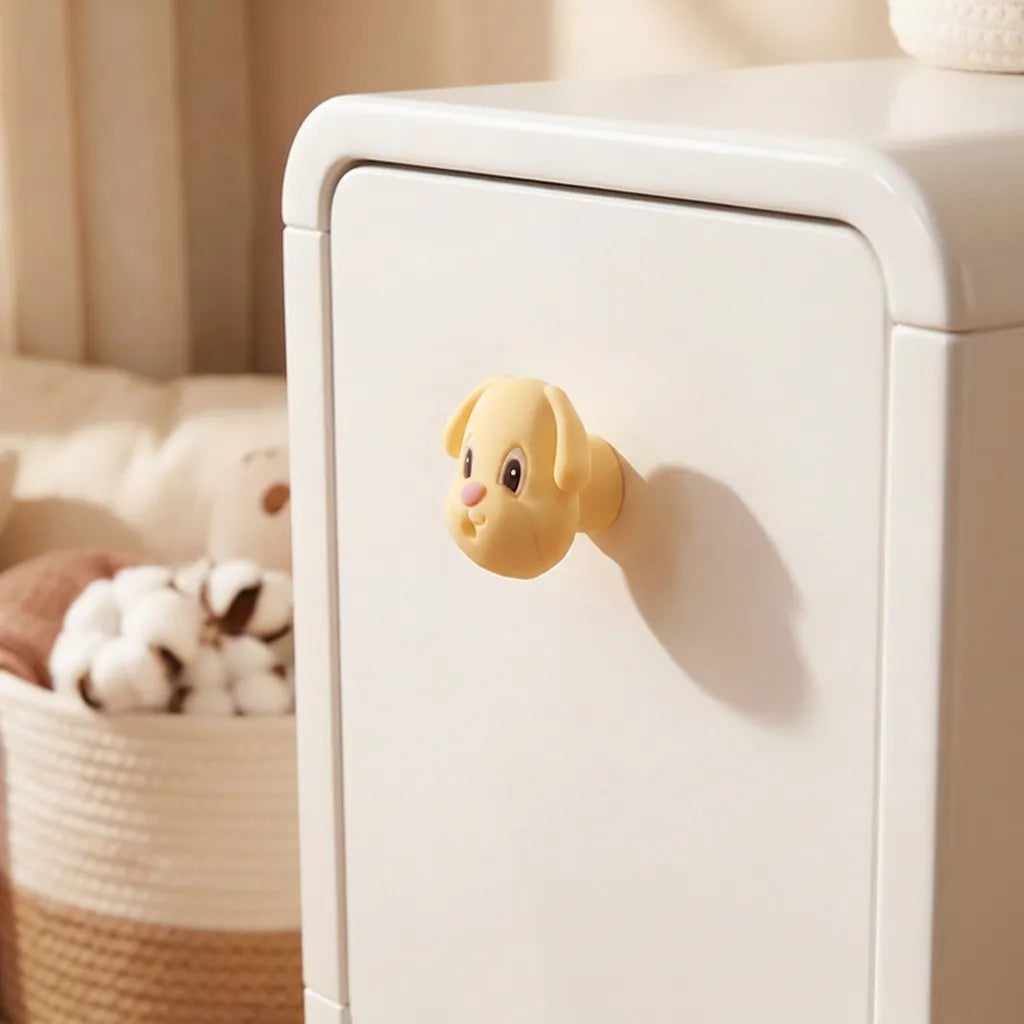 A small yellow dog-shaped drawer pull handle attached to a white cabinet door in a softly lit room, with a woven basket and plush toy blurred in the background.