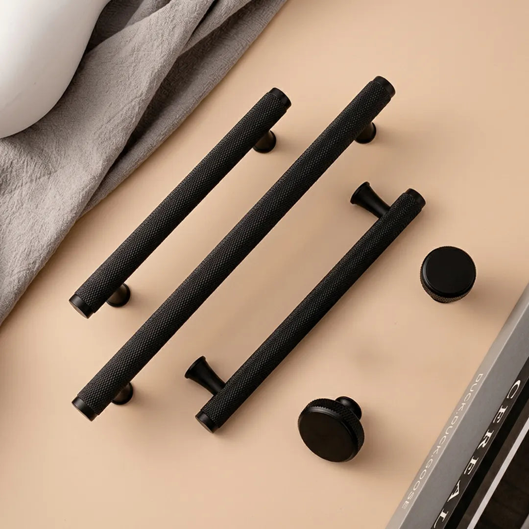 Three long black textured cabinet handles and two round black knobs arranged on a warm beige surface with a folded gray cloth and part of a white object in the background.