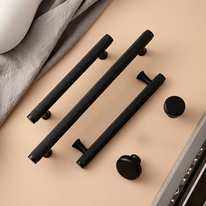 Three long black textured cabinet handles and two round black knobs arranged on a warm beige surface with a folded gray cloth and part of a white object in the background.