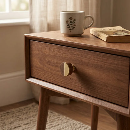 A mid-century modern wooden nightstand with a single drawer and a round brass-and-wood knob, styled in a warm room setting with a mug and book on top.
