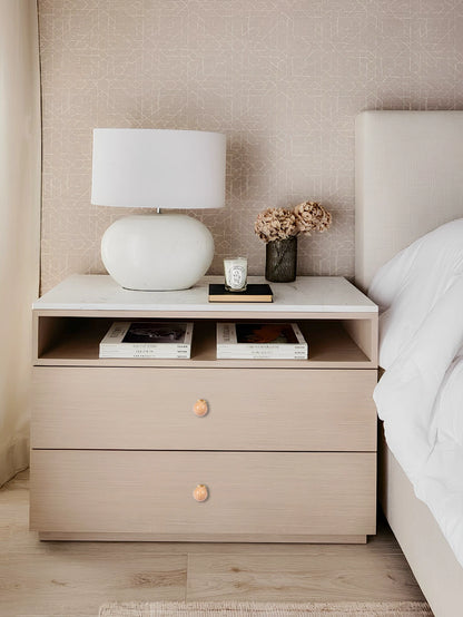 Beige nightstand with a round knob, topped with a lamp, books, and a small plant in a cozy bedroom setting—by Mantara.