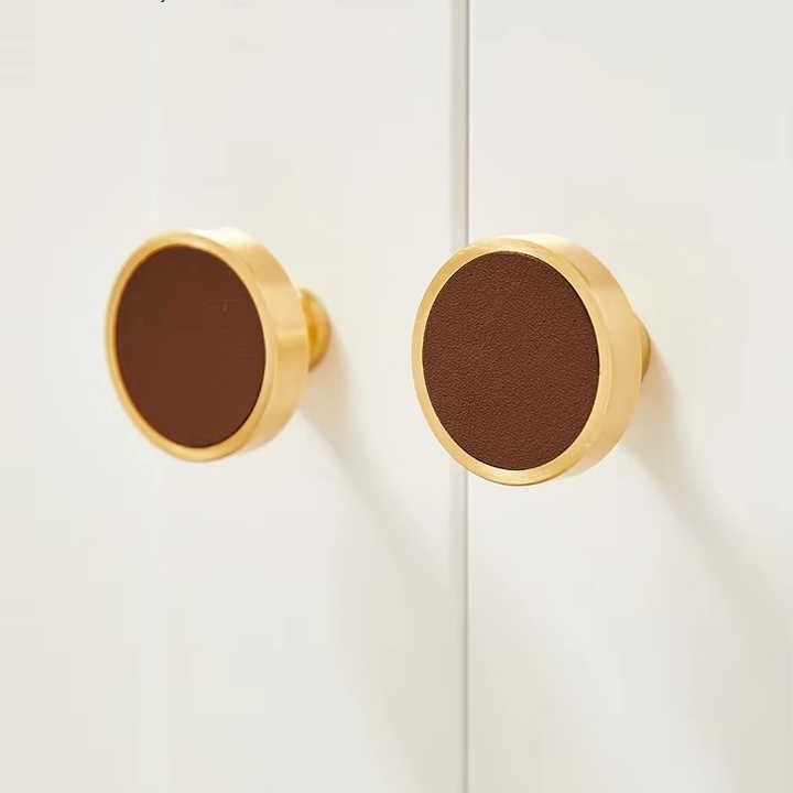 Two round cabinet knobs with brown leather insets and brass edges, mounted on a white surface, styled by Mantara.