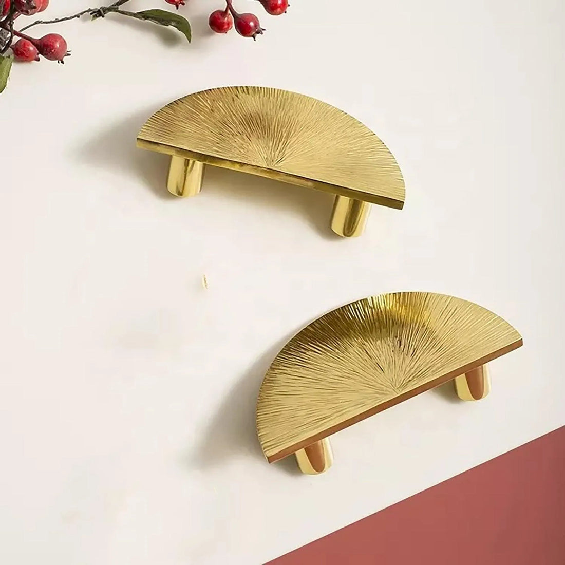 Two semicircular gold cabinet handles with a textured sunburst design are placed on a light surface. Red berries and green leaves are in the background.