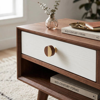 A mid-century modern wooden nightstand with a white drawer front, a round wood-and-brass knob, and open shelving, styled in a bright room with books and a small vase.
