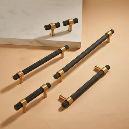 Four black and gold cabinet handles arranged on a warm beige surface, with two smaller T-bar handles resting on a white marble slab in the background.