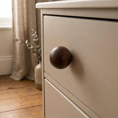 Close-up of a light beige wooden dresser drawer with a round dark brown wooden knob, set in a softly lit room with a curtain, vase, and wood floor.