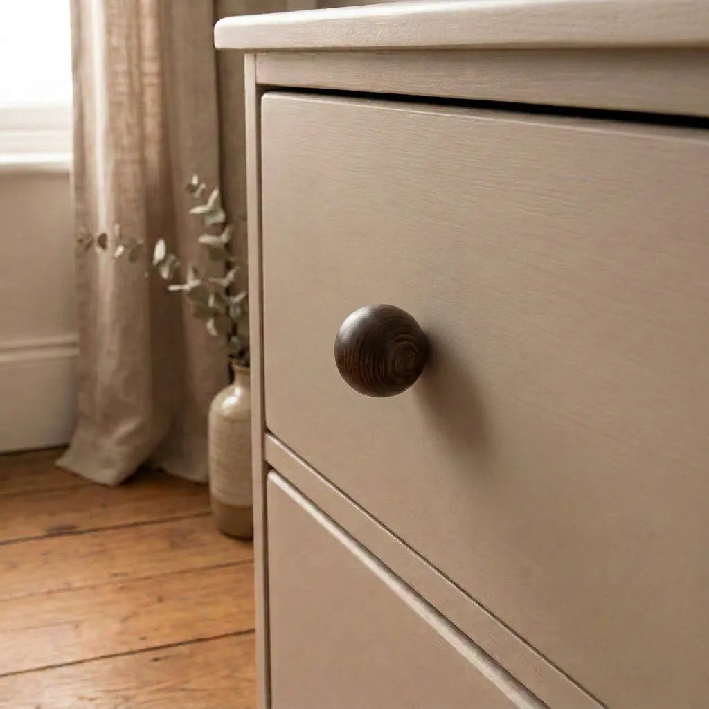 Close-up of a light beige wooden dresser with a dark brown round knob, set in a softly lit room with curtains, a vase, and wood flooring.