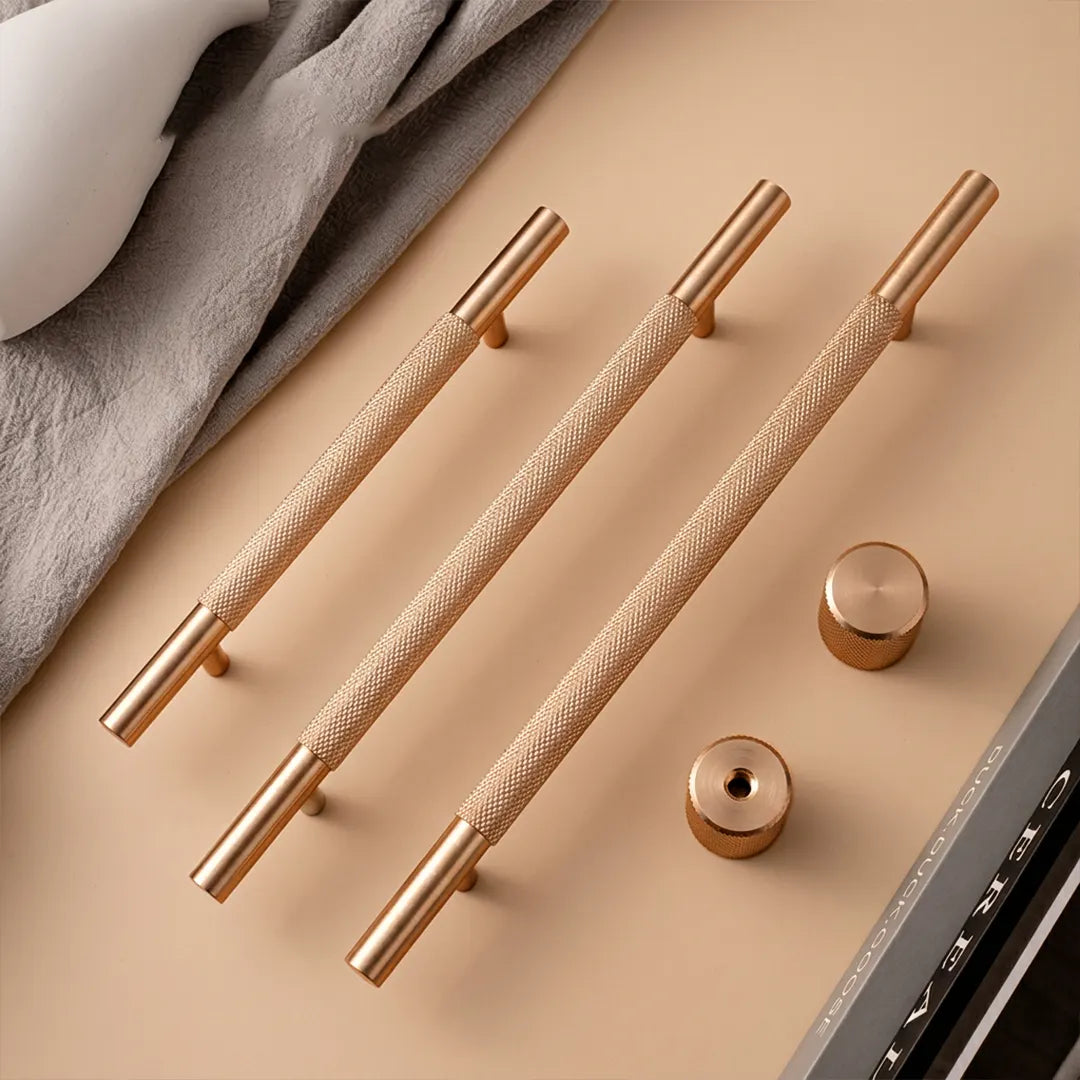 Three elongated brushed brass cabinet handles with textured grips and matching round knobs, arranged on a warm beige surface with a gray fabric and partial laptop edge in the background.