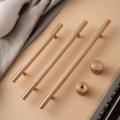 Three elongated brushed brass cabinet handles with textured grips and matching round knobs, arranged on a warm beige surface with a gray fabric and partial laptop edge in the background.