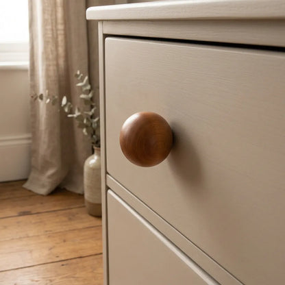Close-up of a light beige painted wooden dresser drawer with a round medium-brown wooden knob, set in a softly lit room with a window, curtain, and wood floor visible in the background.