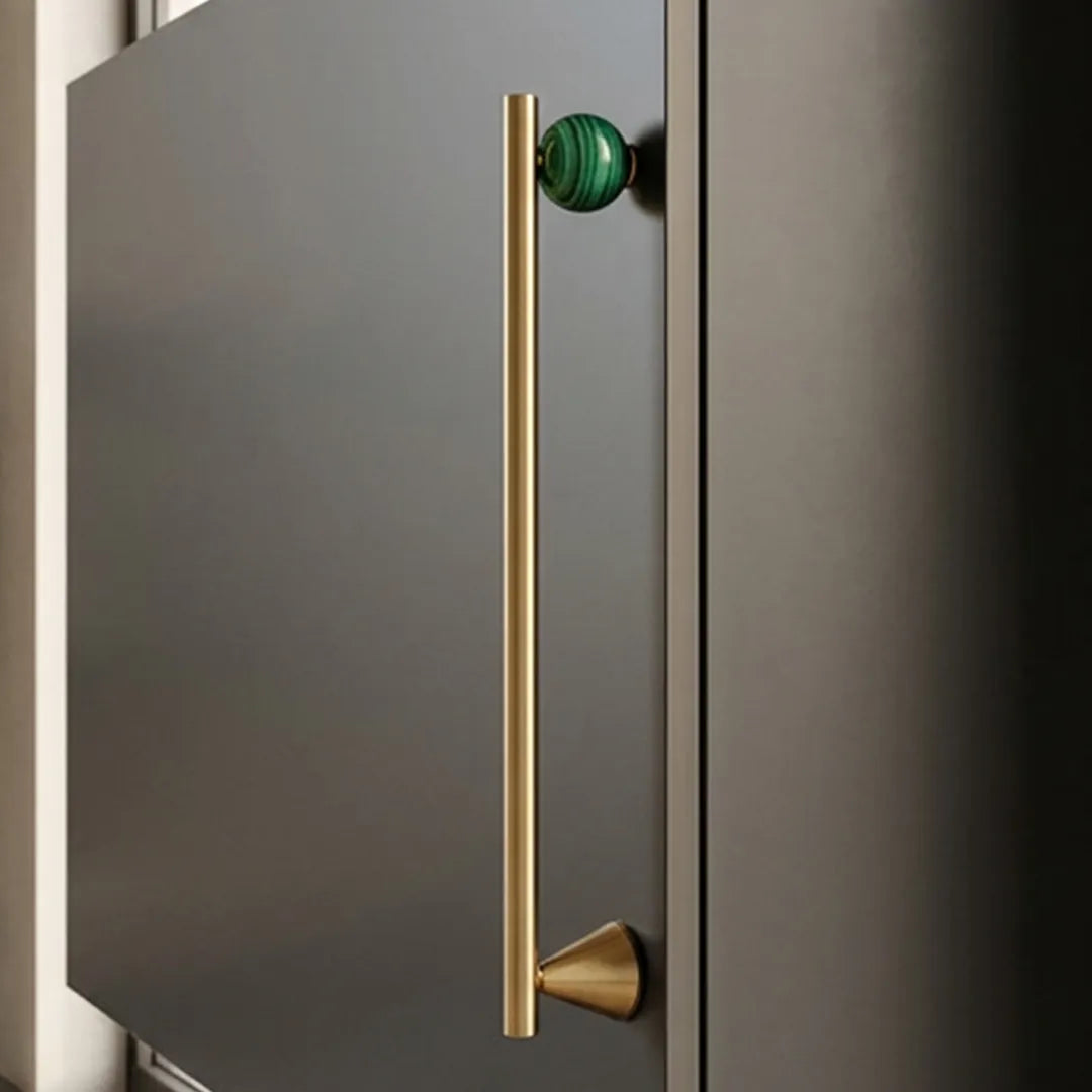 A modern kitchen cabinet featuring a long, slim vertical brass bar handle with a green marble-accented top knob, set against a dark charcoal gray door.