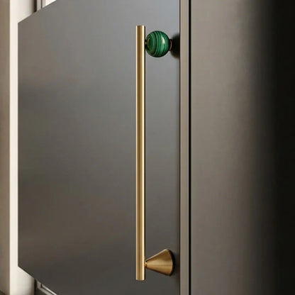 A modern kitchen cabinet featuring a long, slim vertical brass bar handle with a green marble-accented top knob, set against a dark charcoal gray door.