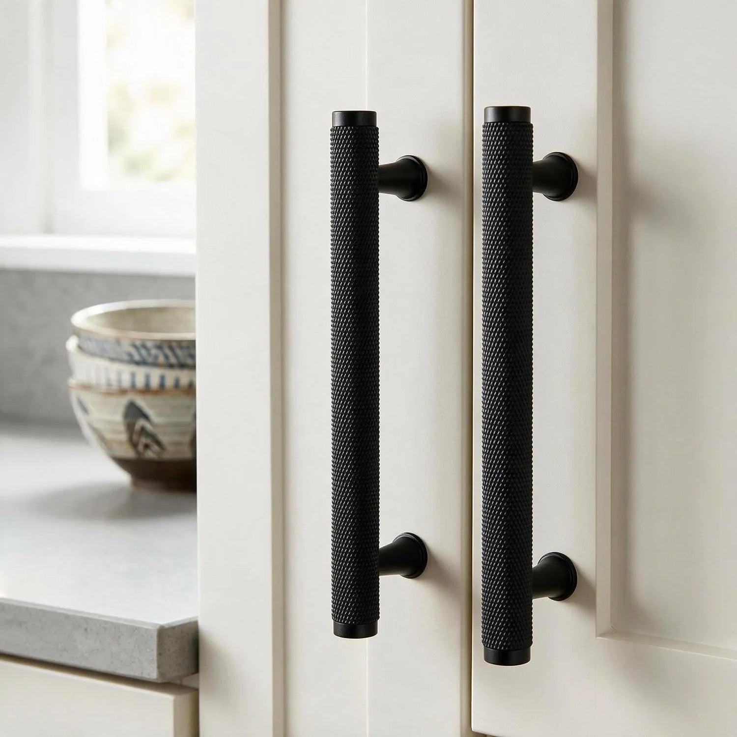 Two long black textured cabinet handles installed on cream kitchen doors, with a countertop and bowls visible in a softly lit kitchen setting.