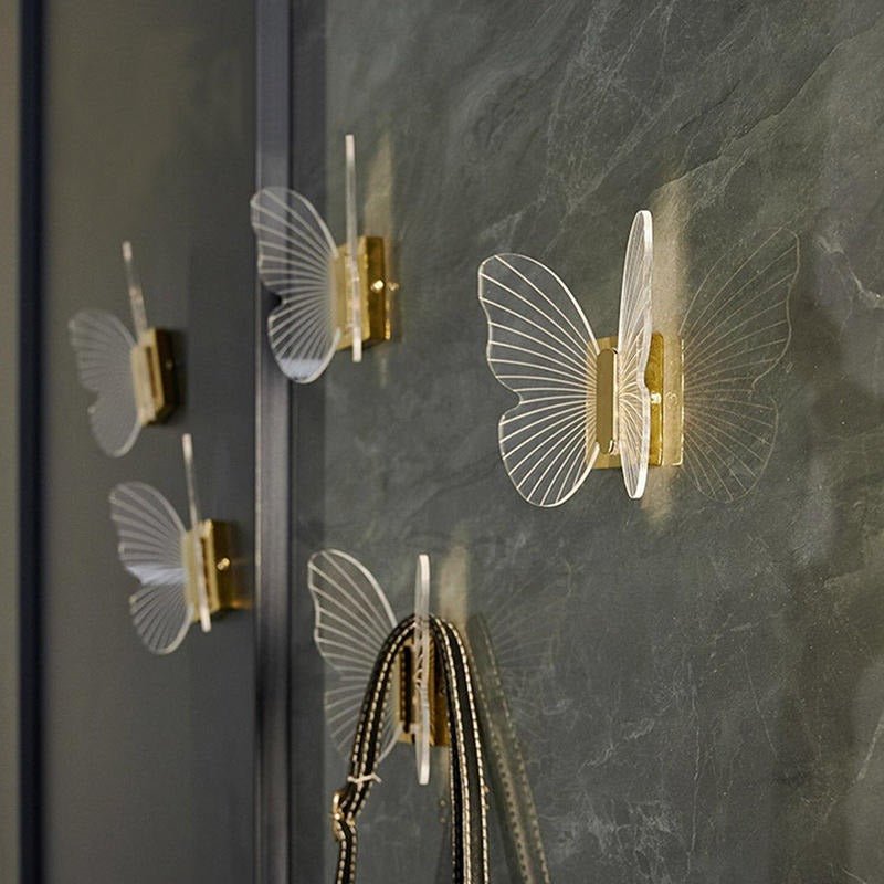 Gold-colored butterfly-shaped wall hooks on a grey wall, with one hook holding a chain—whimsical function by Mantara.